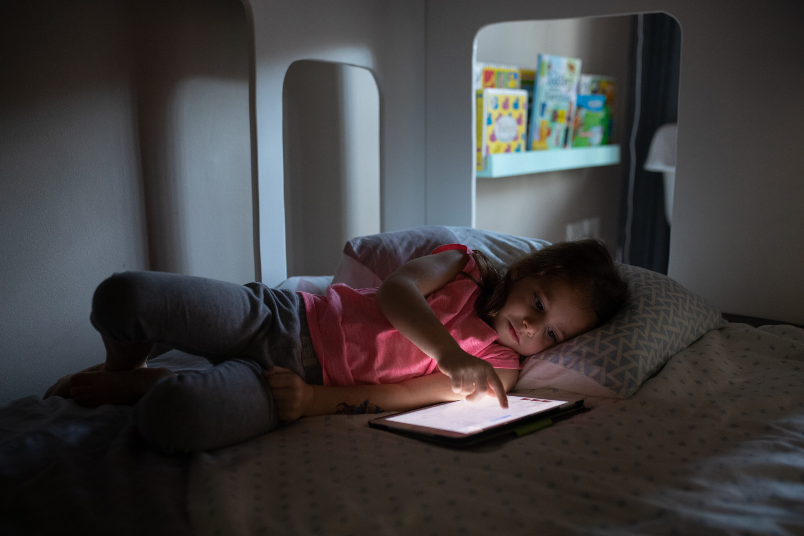 Child lying in bed at night looking at a glowing tablet screen, storybooks visible on a shelf nearby