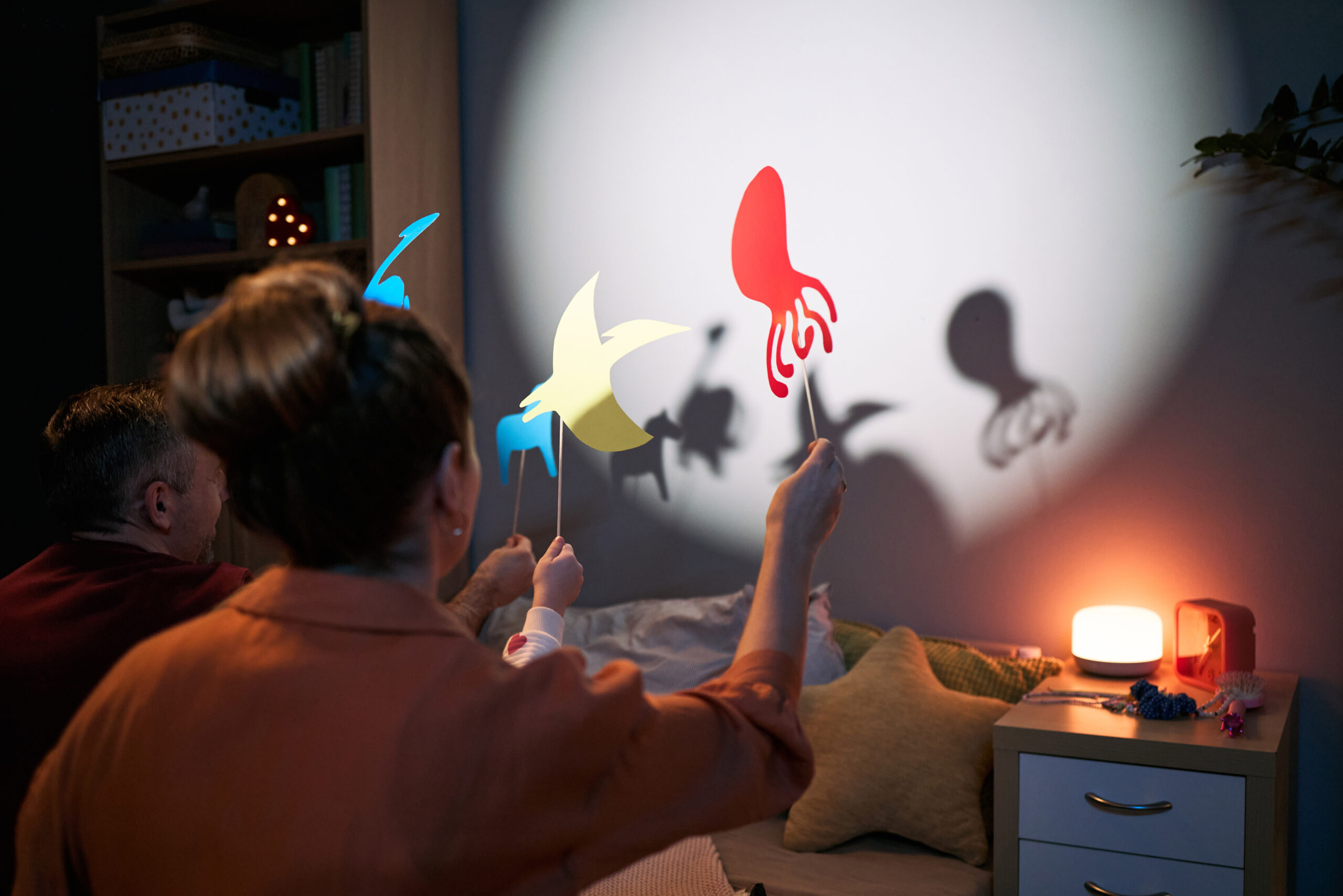 Parents making shadow puppet shapes on the wall with a flashlight for two kids sitting up in bed