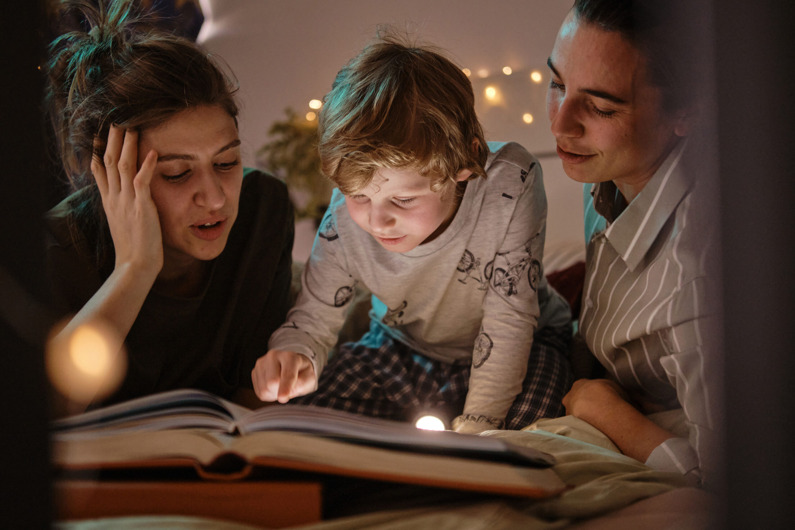 Child pointing at a page in a book while two adults read together at bedtime